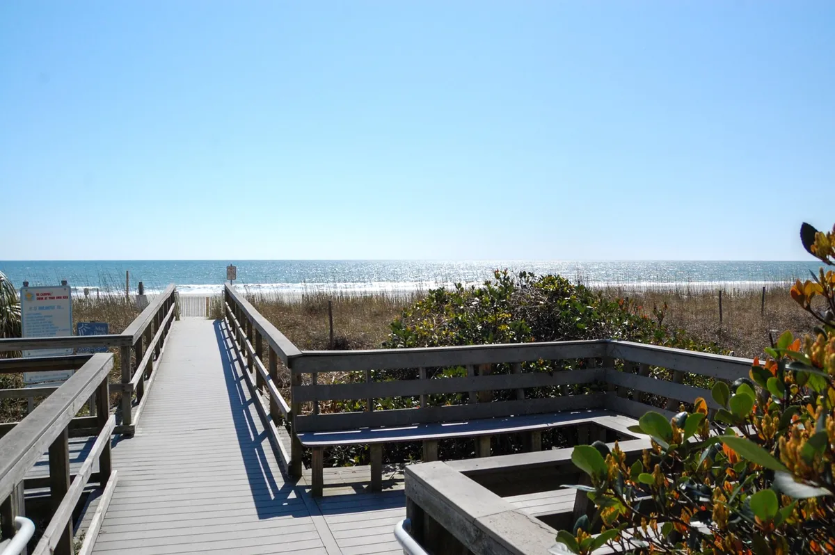 A tranquil wooden walkway leads through lush green vegetation and gentle sand dunes, opening to a stunning ocean view. The early light hints at sunrise, suggesting peaceful morning strolls and direct beach access.