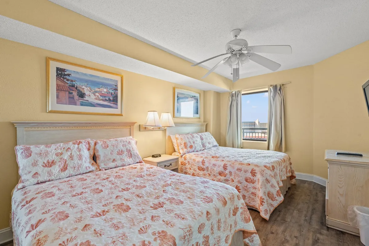 A comfortable bedroom with two beds adorned with seashell-patterned bedspreads, wall art, and a window bringing in natural light. The inviting atmosphere is complemented by decor inspired by the beach, making it ideal for both families and long-term guests.
