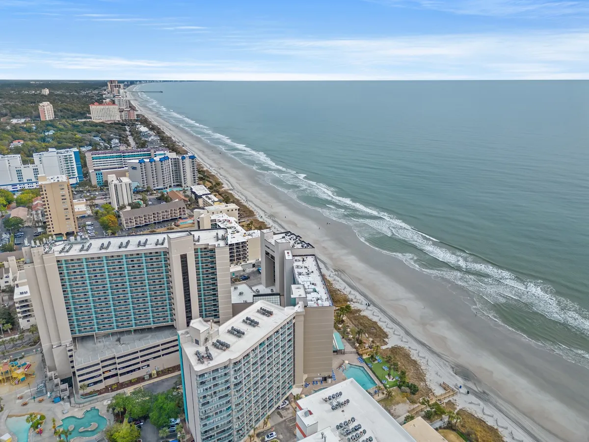 A captivating view from above featuring the sparkling ocean, golden beach, high-rise buildings, and an inviting pool area. The expansive perspective illustrates the interplay between relaxation by the pool and adventures on the beach.