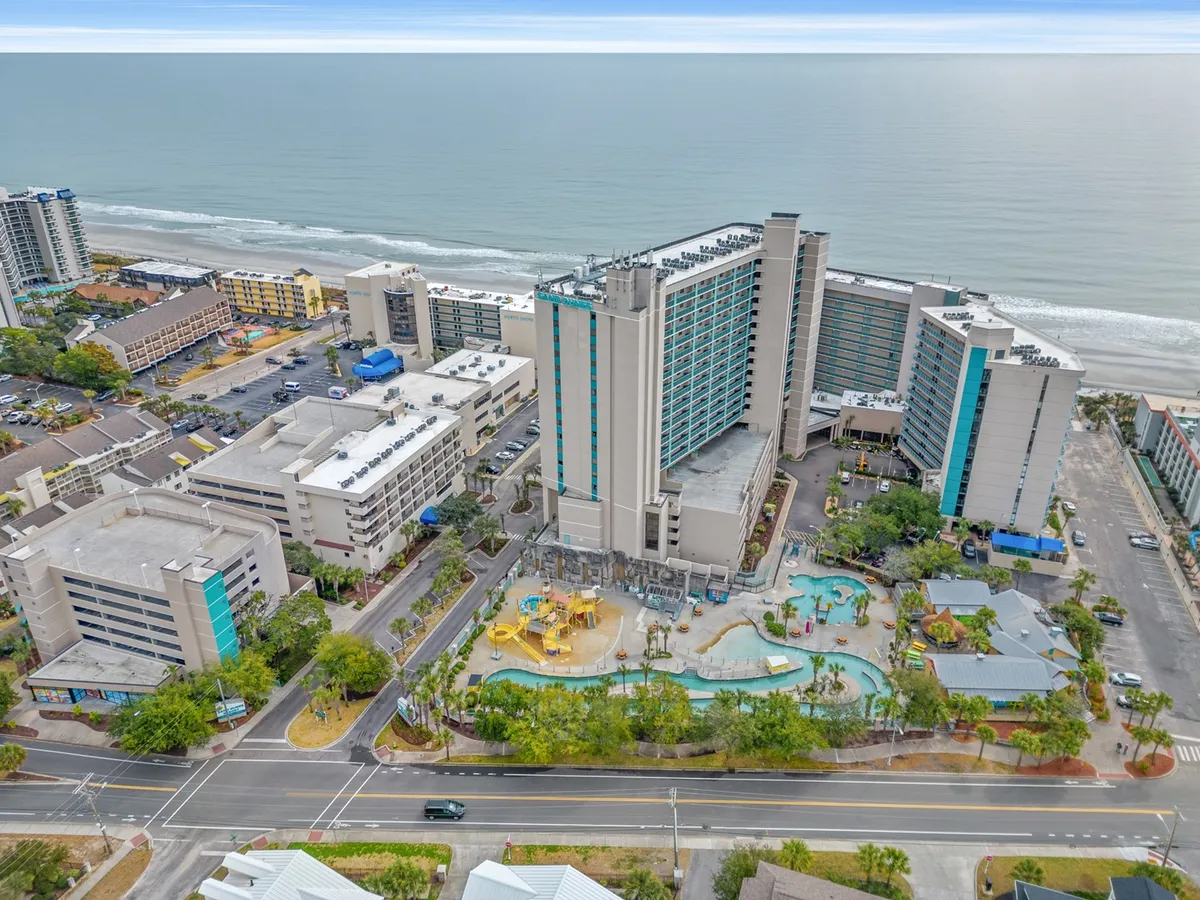 A wide exterior view of the Sand Dunes Resort, featuring a large swimming pool surrounded by loungers, the expansive hotel building, lush greenery, and a sweeping view of the ocean just steps away. The scene captures guests enjoying the pool and the proximity to the beach, with sunlit skies enhancing the joyful atmosphere.