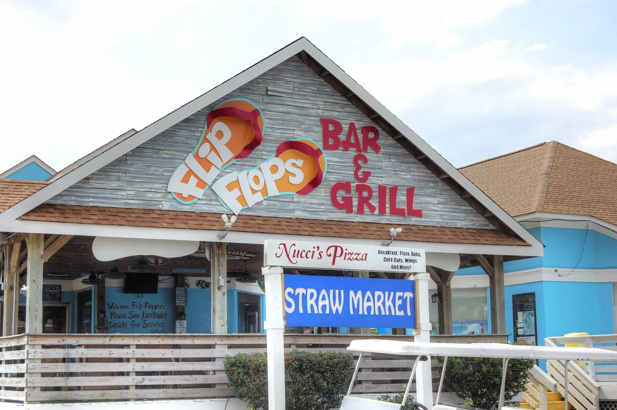 A cluster of vibrant local dining and market signs—Flip Flops Bar & Grill, Nucci's Pizza, and Straw Market—creates a lively scene perfect for casual outings. The area is filled with promise of socializing, local flavors, and year-round entertainment.