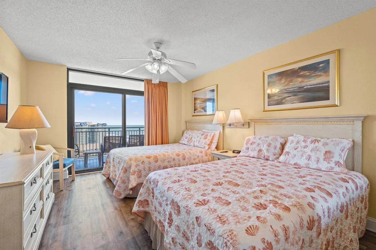 A tranquil bedroom featuring two beds, an ocean view window, and a nightstand with a lamp. The warm, natural light filters in, creating a serene and cozy atmosphere perfect for unwinding after a day at the beach.