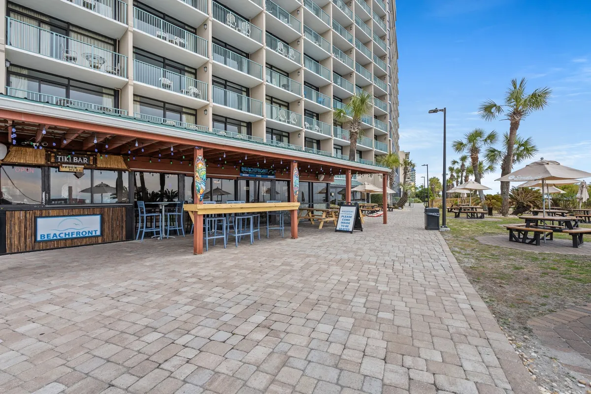This vibrant exterior scene focuses on a beachfront Tiki Bar, palm trees, and an inviting outdoor seating area, with beach access visible nearby. The mood is casual and festive, suggesting opportunities for casual meals and socializing steps from the sand.