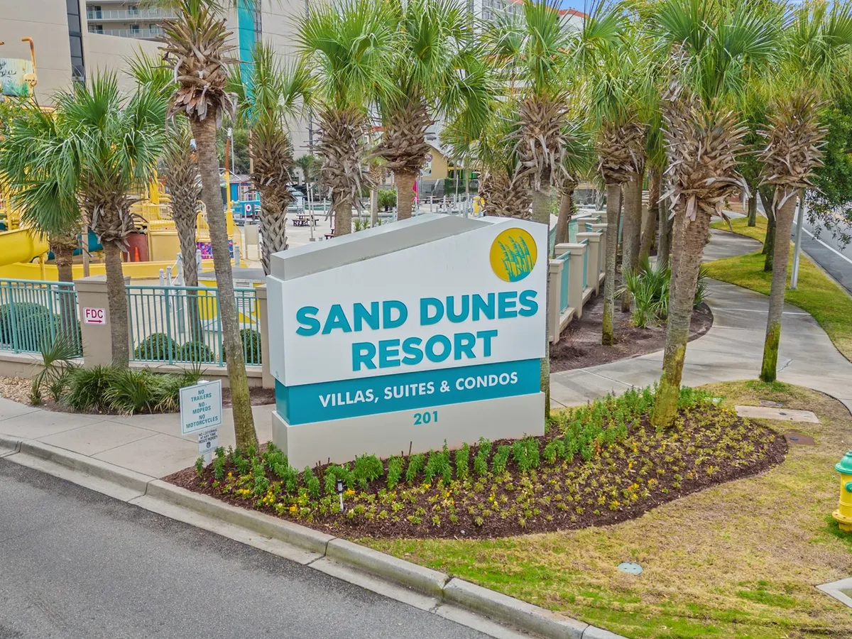 A welcoming view of the Sand Dunes Resort exterior, featuring the signature resort sign, lush palm trees, and a neatly landscaped garden. The scene is set during a bright, inviting moment, hinting at the warmth and hospitality guests can expect during their stay.