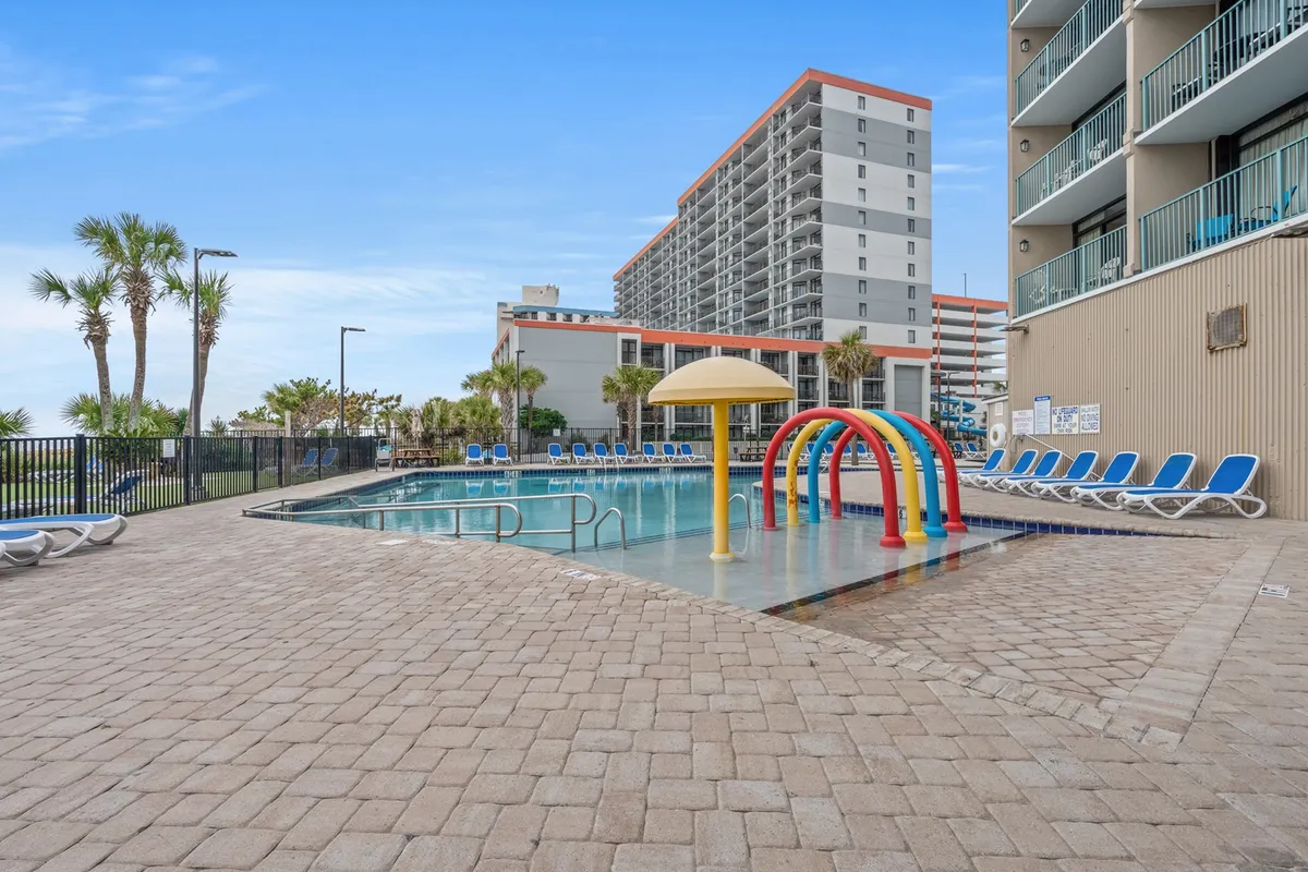 A cheerful outdoor pool area surrounded by palm trees and sun loungers, featuring a colorful water play zone designed for children and families. The image radiates joy, with families gathered poolside and kids splashing in the water under the warm sun.