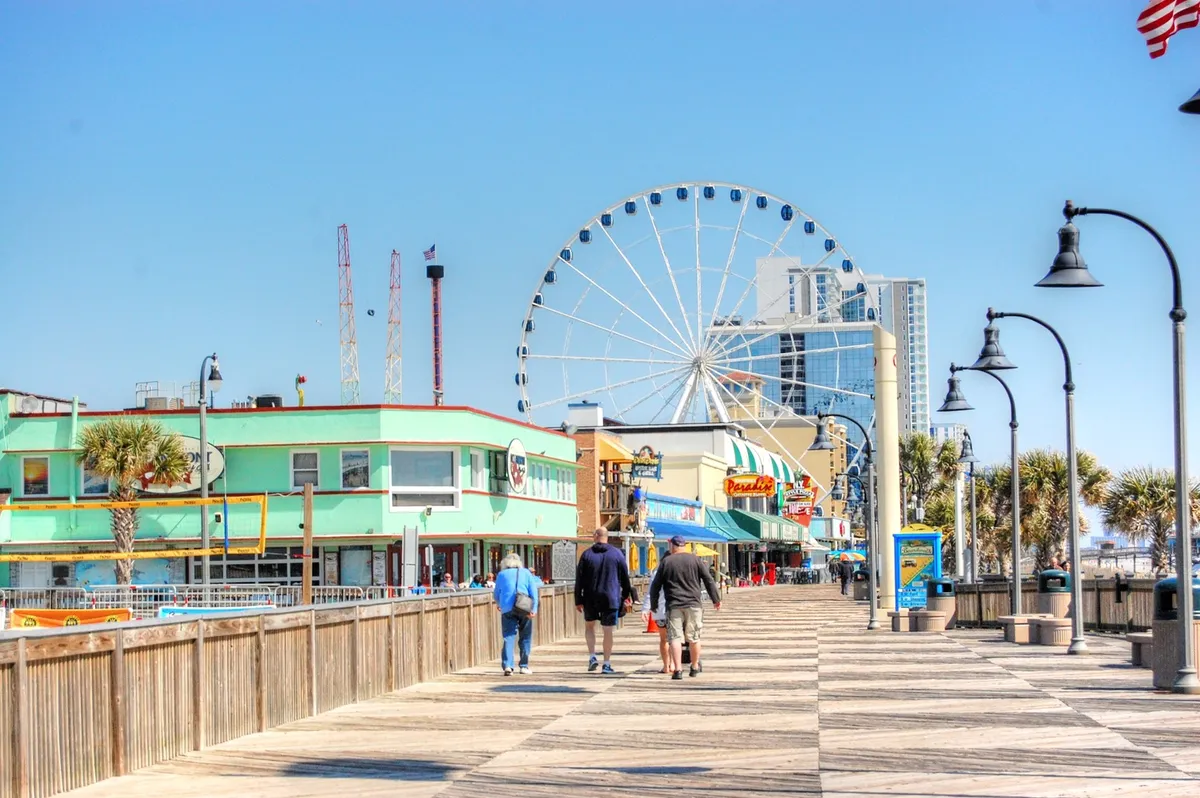 A vibrant shot of Myrtle Beach’s lively boardwalk, complete with a towering ferris wheel, colorful storefronts, and families exploring the shops and enjoying the coastal breeze. The scene is bustling with energy, evoking the fun and excitement of local attractions within close reach of the resort.