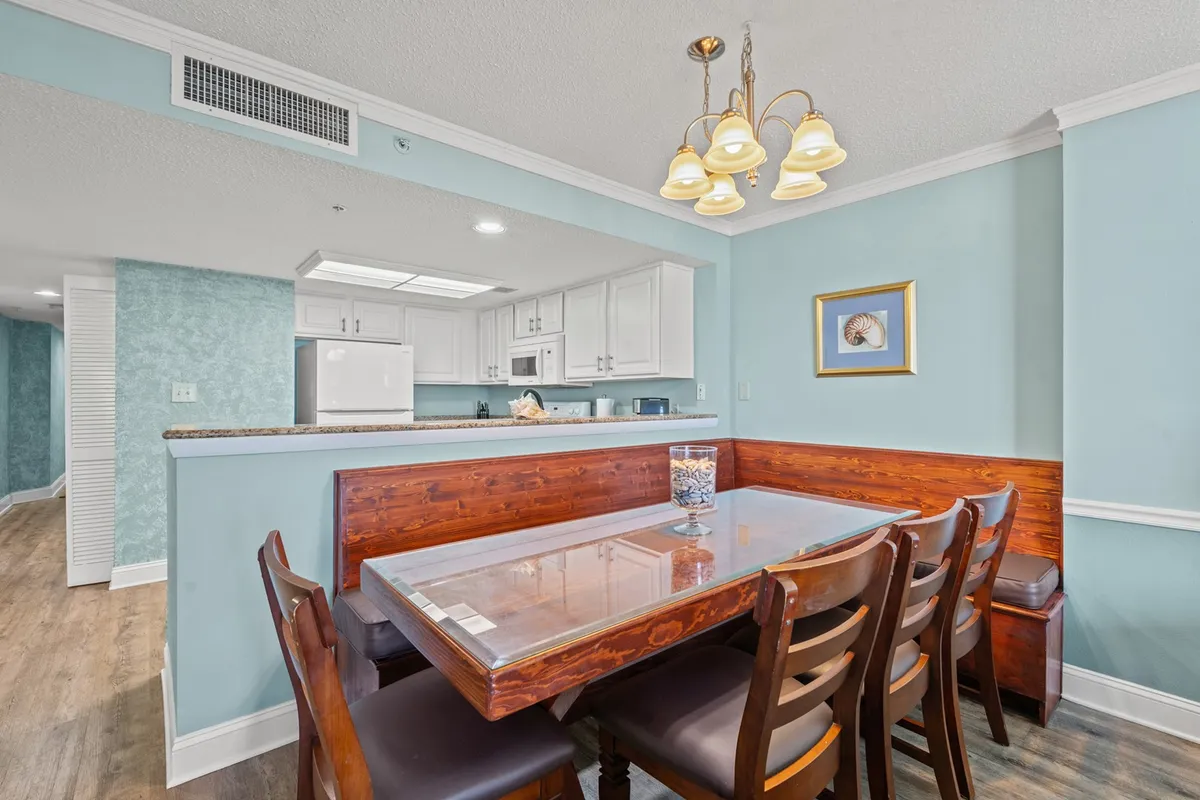 A warm dining area with a central table, elegant chandelier, and a view into the adjacent kitchen. Tasteful wall art and comfortable seating create an inviting atmosphere for shared meals and celebrations.
