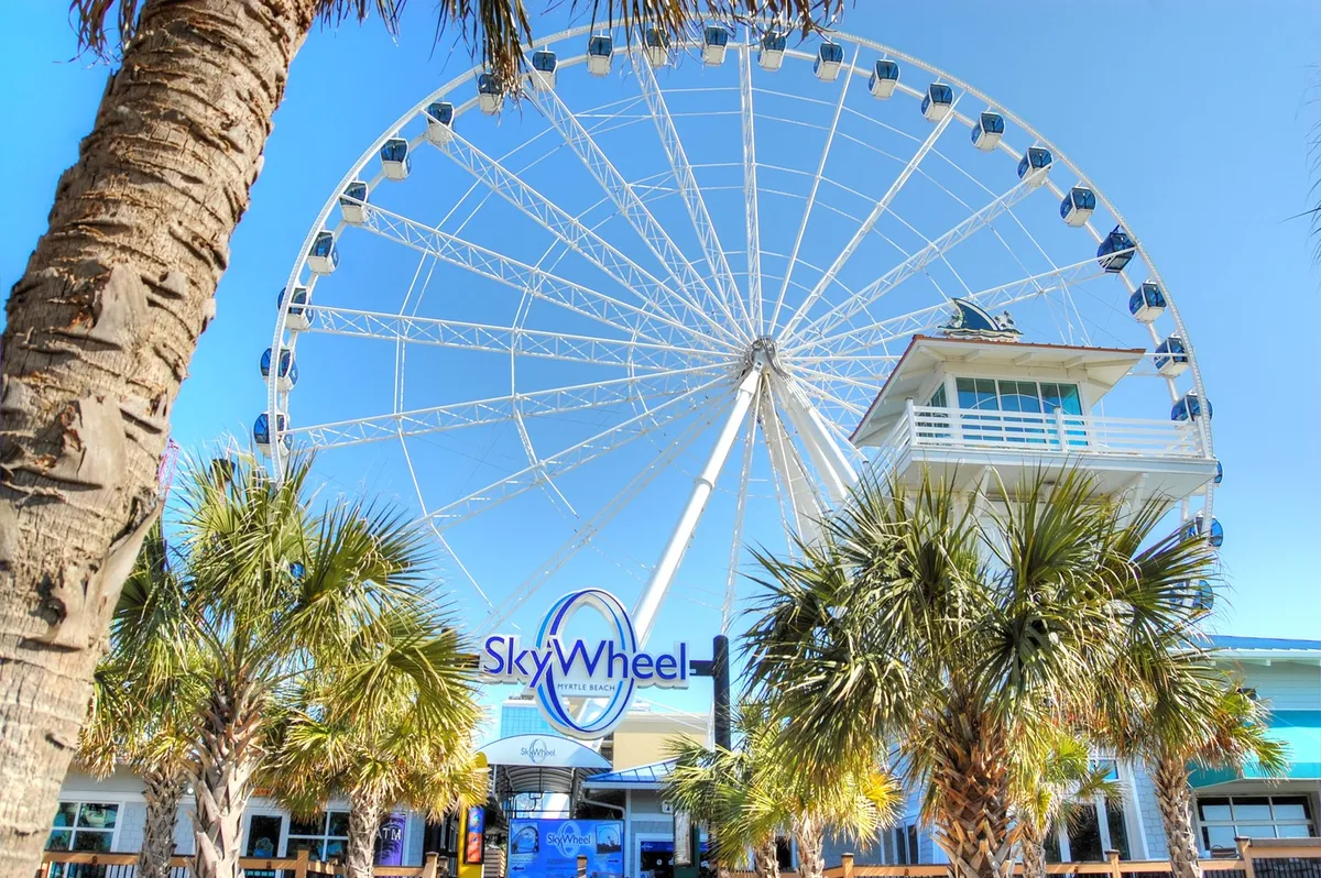 An eye-catching exterior scene of the iconic Myrtle Beach SkyWheel towering above palm trees against a clear blue sky. The setting is bustling and energetic, inviting viewers to imagine the thrill and memorable moments awaiting at this popular local attraction.