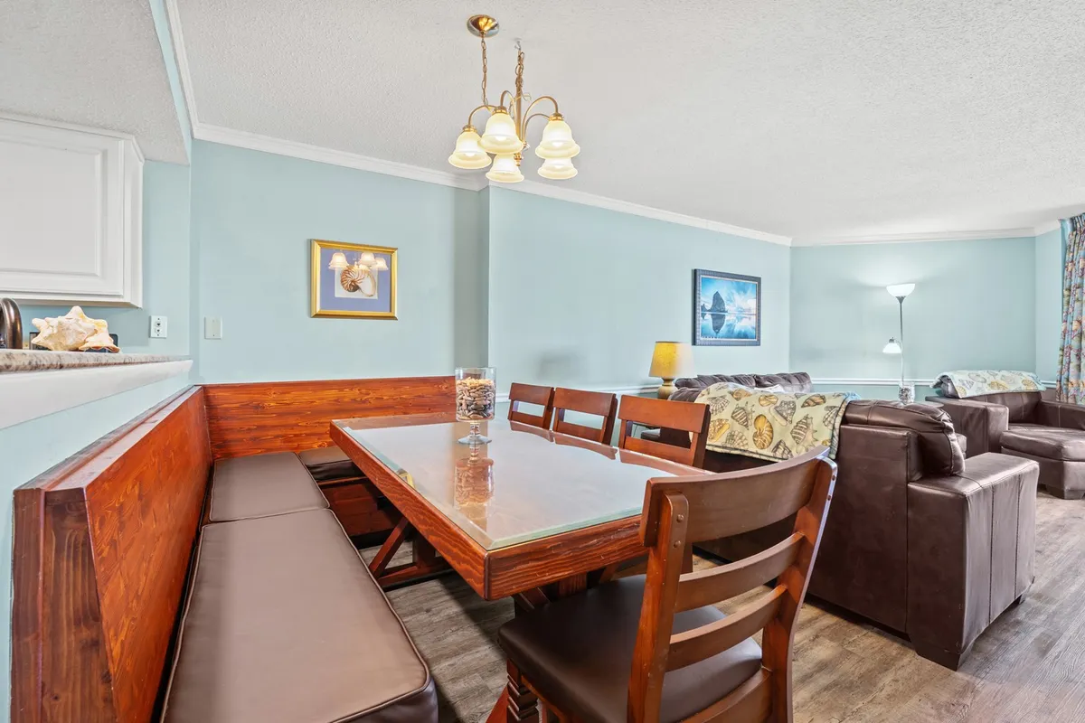 A warmly lit dining area takes center stage in this image, featuring a large dining table surrounded by chairs, stylish wall art, and a glimpse of the adjacent living space. The setup suggests a space ideal for family meals and group celebrations.