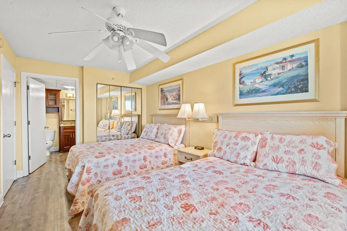 A spacious bedroom with two queen beds, framed artwork, soft lighting from table lamps, and a mirrored closet providing ample storage. The room conveys a sense of spaciousness and modern comfort for families or groups.