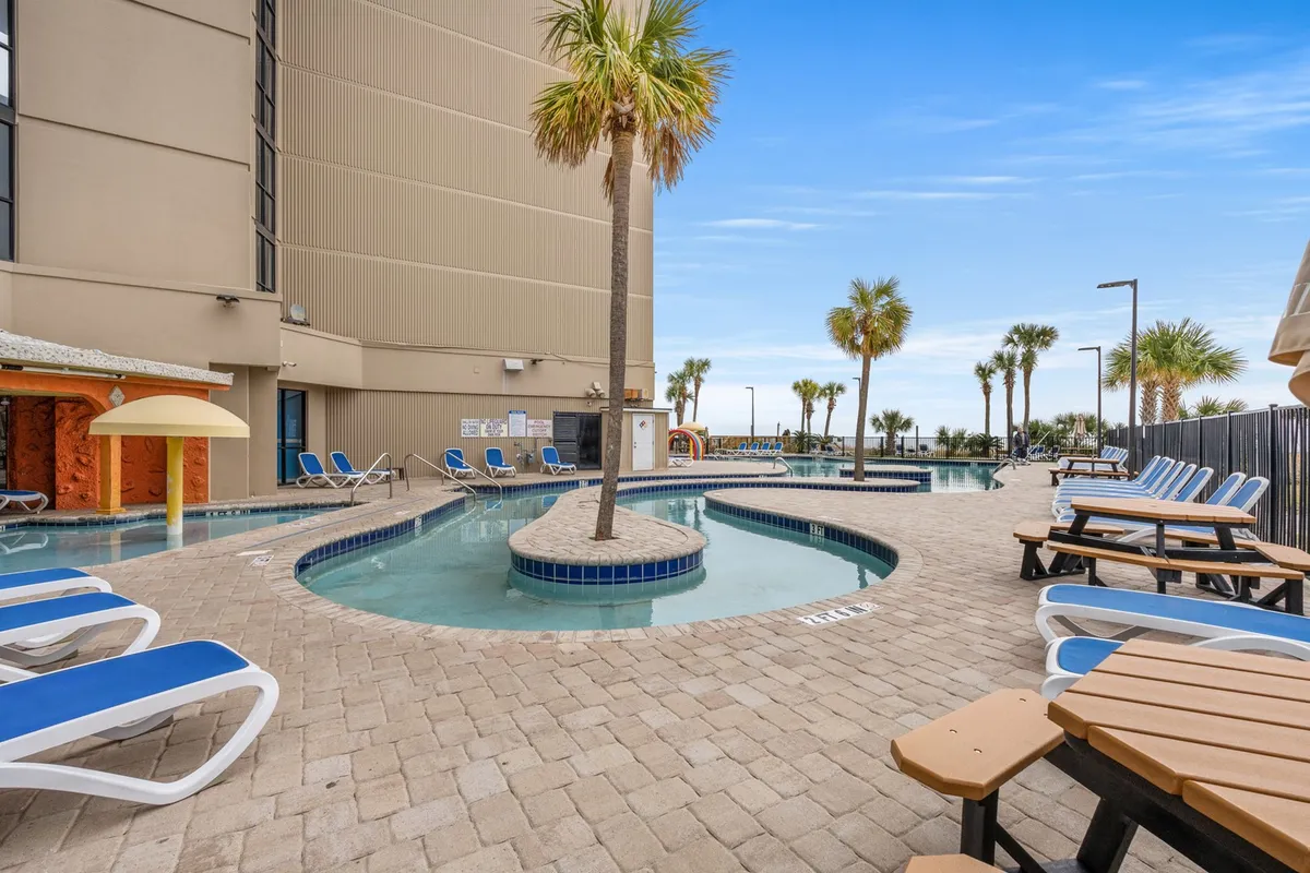 This image captures an outdoor swimming pool surrounded by lounge chairs, palm trees, and water features, evoking a leisurely, carefree mood ideal for sunbathing, swimming, and social gatherings.
